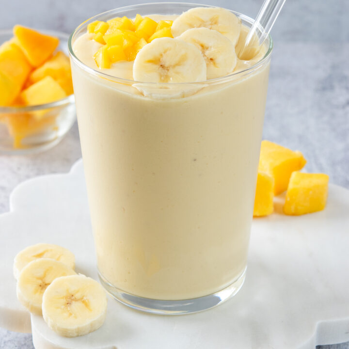 A homemade mango banana smoothie garnished with a few banana slices and chopped fresh mango. The smoothie is in a glass with a glass straw that has a white flower on it.