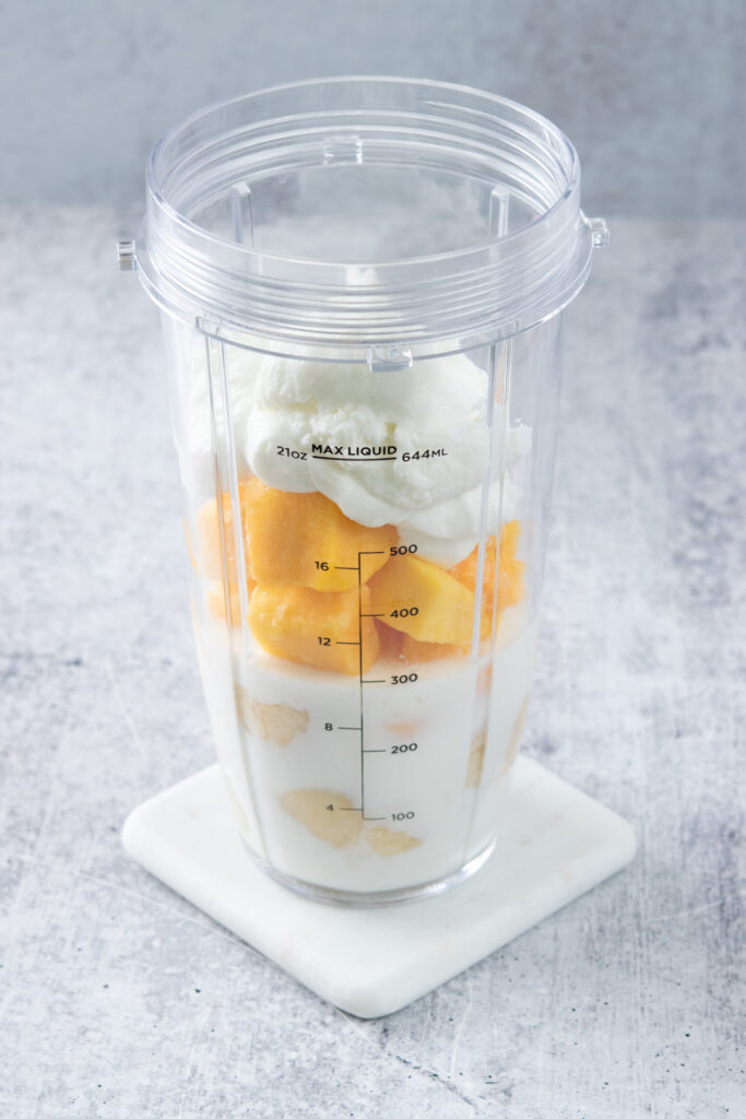 Four ingredients needed to make a mango banana smoothie layered in a personal blender cup in this order: milk, banana, mango, yogurt.