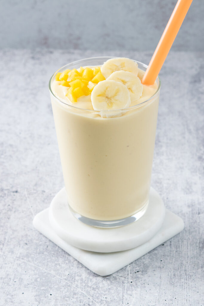 A super creamy mango banana smoothie topped with chopped mango and banana slices, in a glass that has an orange straw in it.