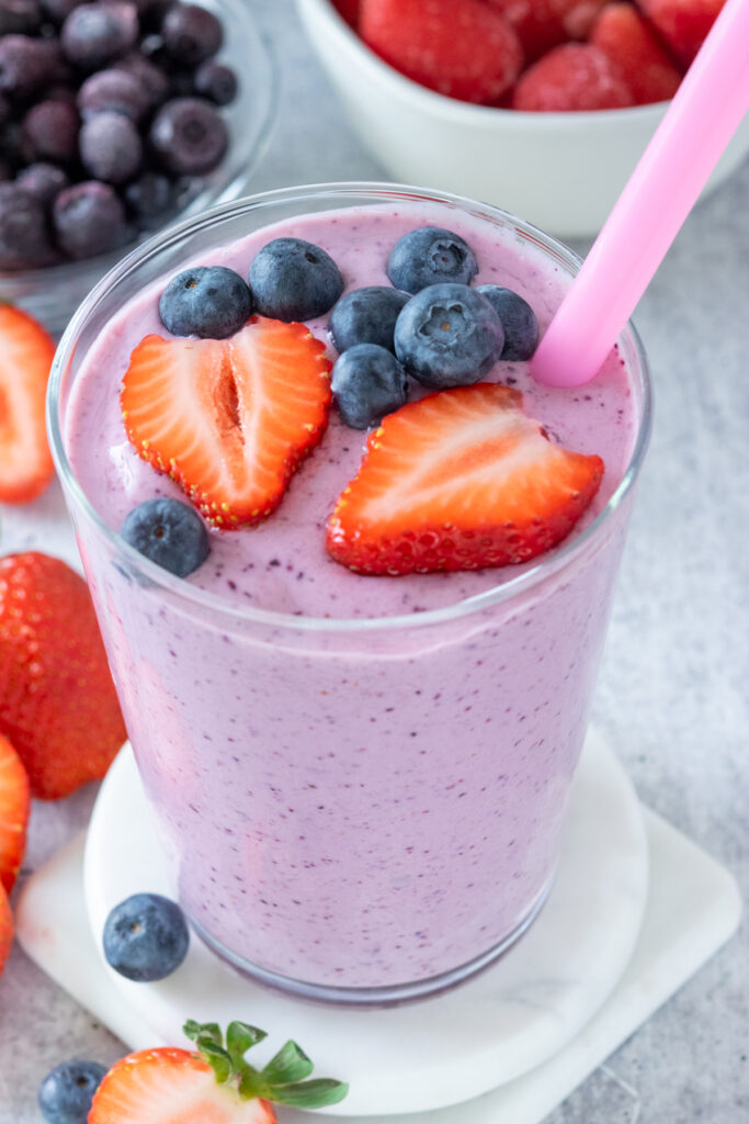 A strawberry blueberry smoothie that has a pink straw in the cup and has some fresh strawberries and blueberries on top. The smoothie is in a glass showing its purple color with flecks of blueberry in it. The smoothie is stacked on top of two white coasters and is next to a small bowl filled with frozen blueberries and another one filled with frozen strawberries.