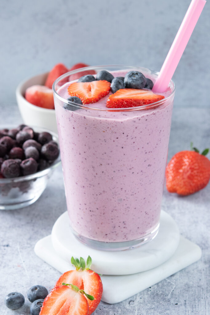 A glass full of a light purple blended strawberry blueberry smoothie that has a pink straw in it and fresh berries on top. The drink is stacked on two white coasters that have strawberry halves and a few blueberries next to them.