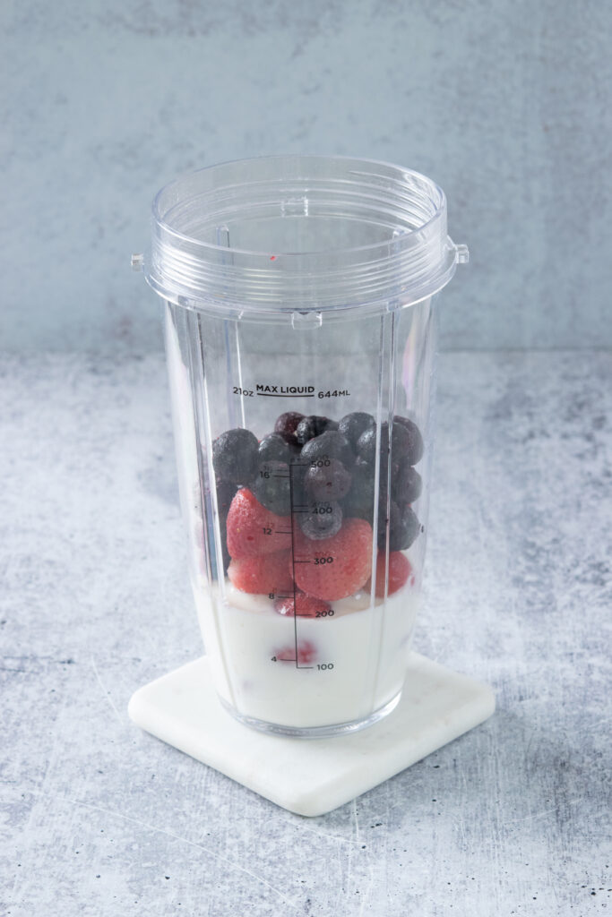 A blender cup that has milk, frozen strawberries and frozen blueberries in it.