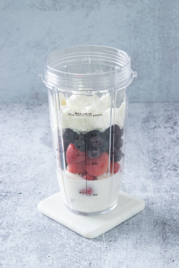 A blender cup that has milk, frozen strawberries, frozen blueberries and Greek yogurt in it.