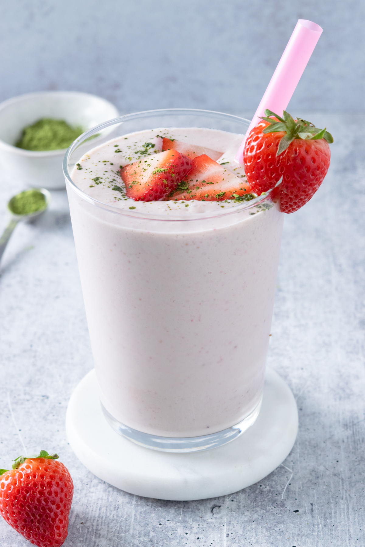 A strawberry matcha smoothie with sliced strawberries and matcha sprinkled on top. The smoothie has a pink straw in it and there's a strawberry next to the cup and a small bowl of matcha powder in the background.